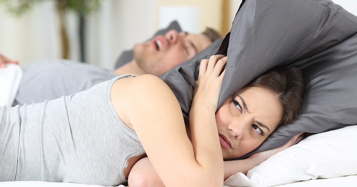 woman covering ears with pillow in bed while man snores